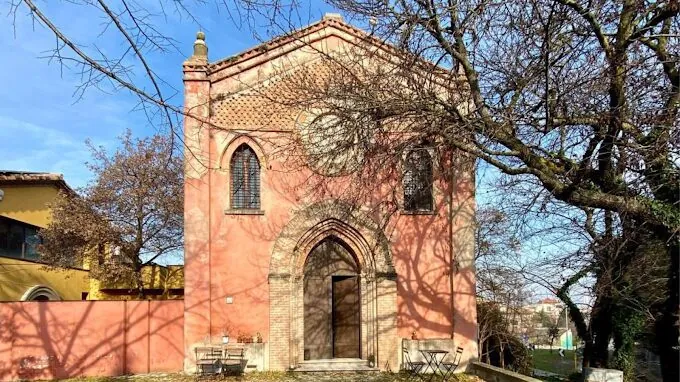 La facciata in mattoni rosa della chiesa di Sant'Andrea dell'Ausa, con il rosone e il portale ad arco, tra gli alberi